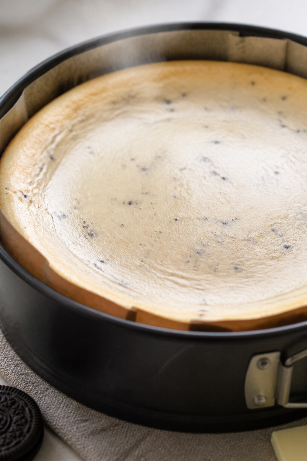 Close-up detail: A just-baked High Protein White Chocolate Oreo Cheesecake cooling on the counter, s