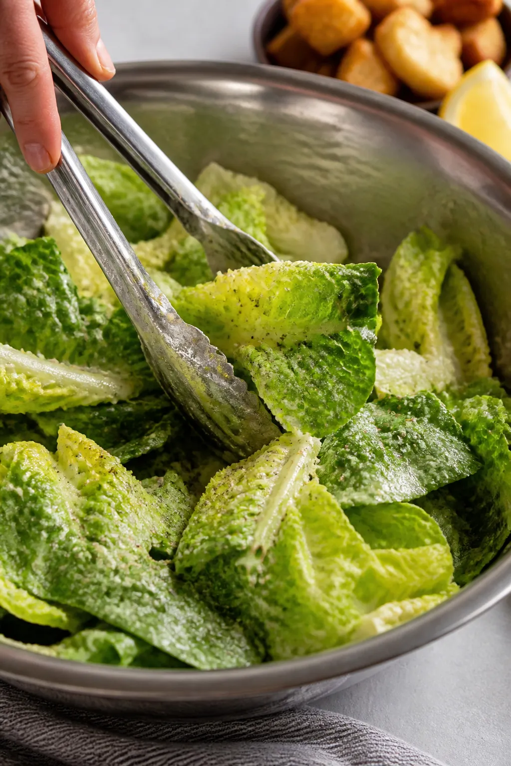 Close-up detail: Glossy Caesar-dressed romaine leaves being tossed in a large stainless bowl with to