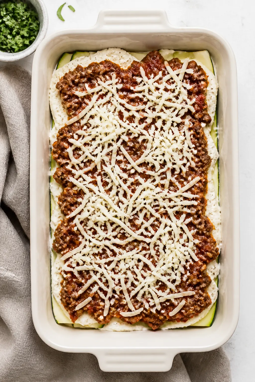 Overhead assembly shot, tasty top view: A 9x13 baking dish mid-assembly of keto ground beef zucchini