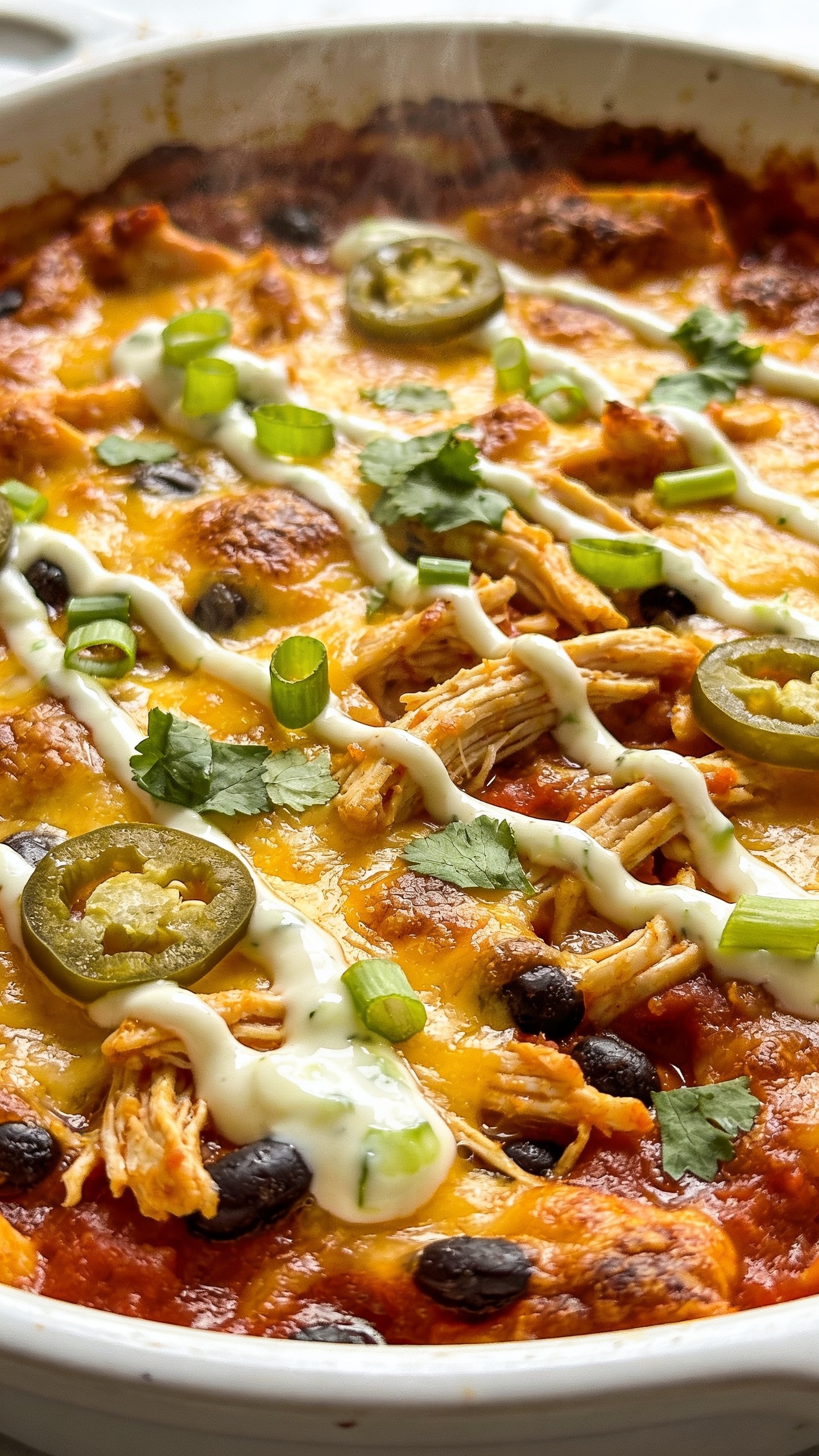 Ultra-close hero shot of a freshly baked high-protein chicken taco casserole in a white ceramic baking dish, filling 90–95% of the frame. Top is deeply golden with bubbly, melted cheddar-jack cheese, crispy browned edges, and pockets of juicy shredded chicken and black beans peeking through a zesty tomato-chile sauce. Visible toppings: finely sliced green onions, chopped cilantro, and a few thin jalapeño rings scattered naturally. A light drizzle of creamy Greek-yogurt-lime sauce creates glossy ribbons across the surface. No text, no hands. Bright natural side-lighting, shallow depth of field, premium food-blog styling, steam gently rising.