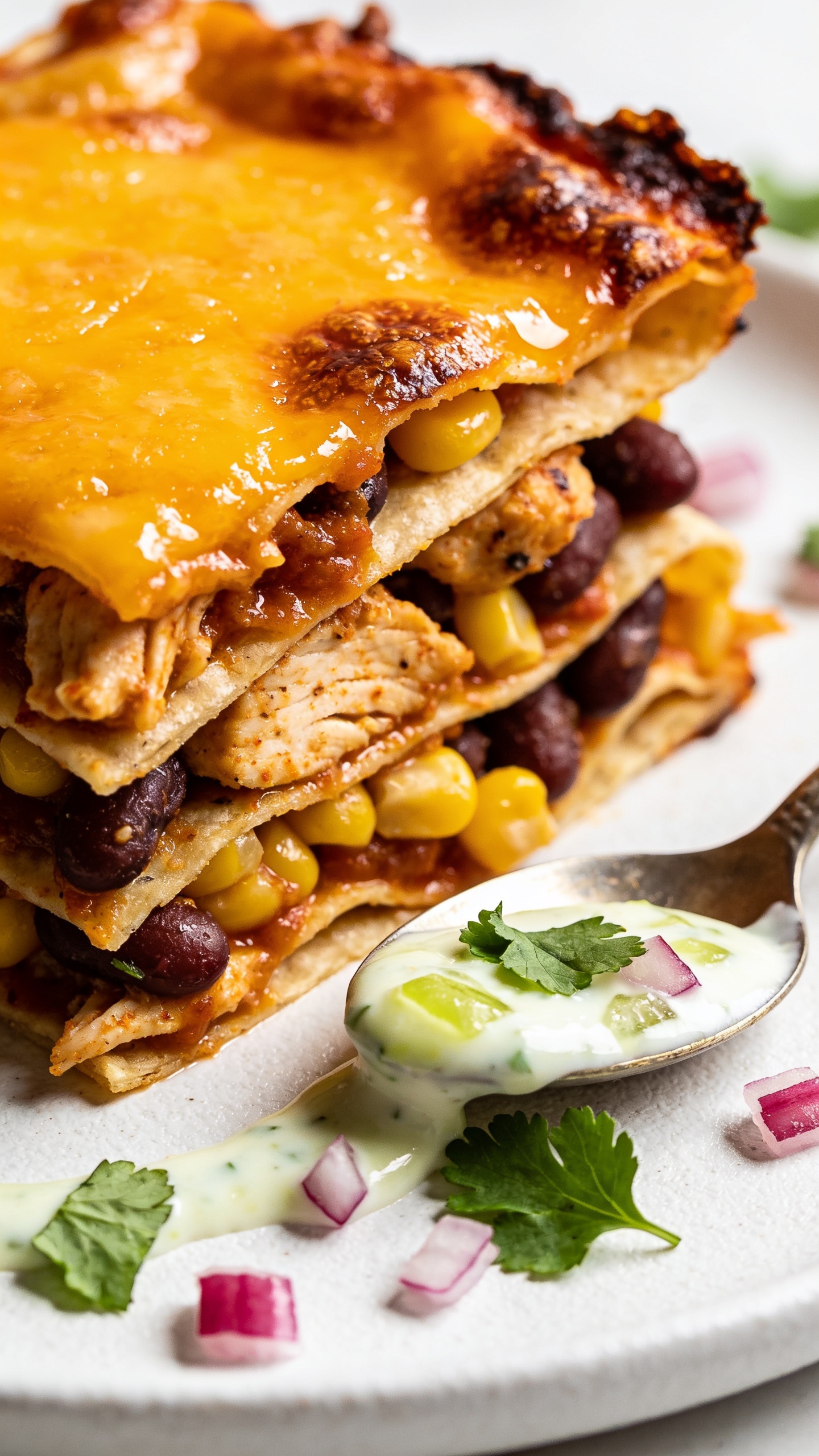 Extreme close-up angle from the corner slice of the same casserole, plated on a matte white plate but cropped so the food fills 90–95% of the frame. Emphasize stacked layers: saucy tortillas, seasoned chicken, beans, and corn, sealed with a glossy, deeply melted cheese cap and crispy browned edges. A delicate spoon-drizzle of Greek-yogurt-lime sauce pools slightly along the edge, with a few fresh cilantro leaves and micro-diced red onion for contrast. Bright natural light, shallow depth of field, rich textures and vibrant colors consistent with the first two images.