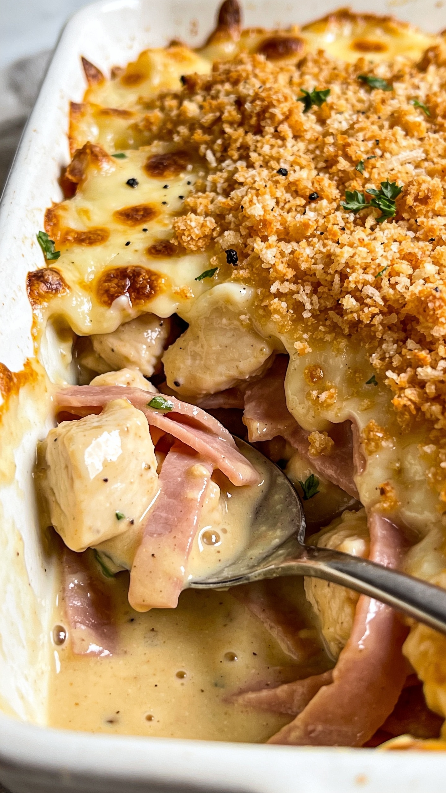 Extreme close-up from a low angle into the corner of the same casserole dish, showcasing a spooned-back section revealing the interior: tender chunks of chicken and thin ribbons of ham coated in a velvety, glossy Dijon-garlic sauce, topped by a thick layer of crunchy, golden-brown panko-parmesan crumbs and melted Swiss browned in spots. A few flecks of black pepper and finely chopped parsley add contrast. Tiny sauce bubbles and cheese blisters catch the light. Bright natural lighting, shallow depth of field with focus on the creamy, saucy interior meeting the crisp topping, minimal neutral background blur, food occupying 90â95% of the frame. No text, no hands.