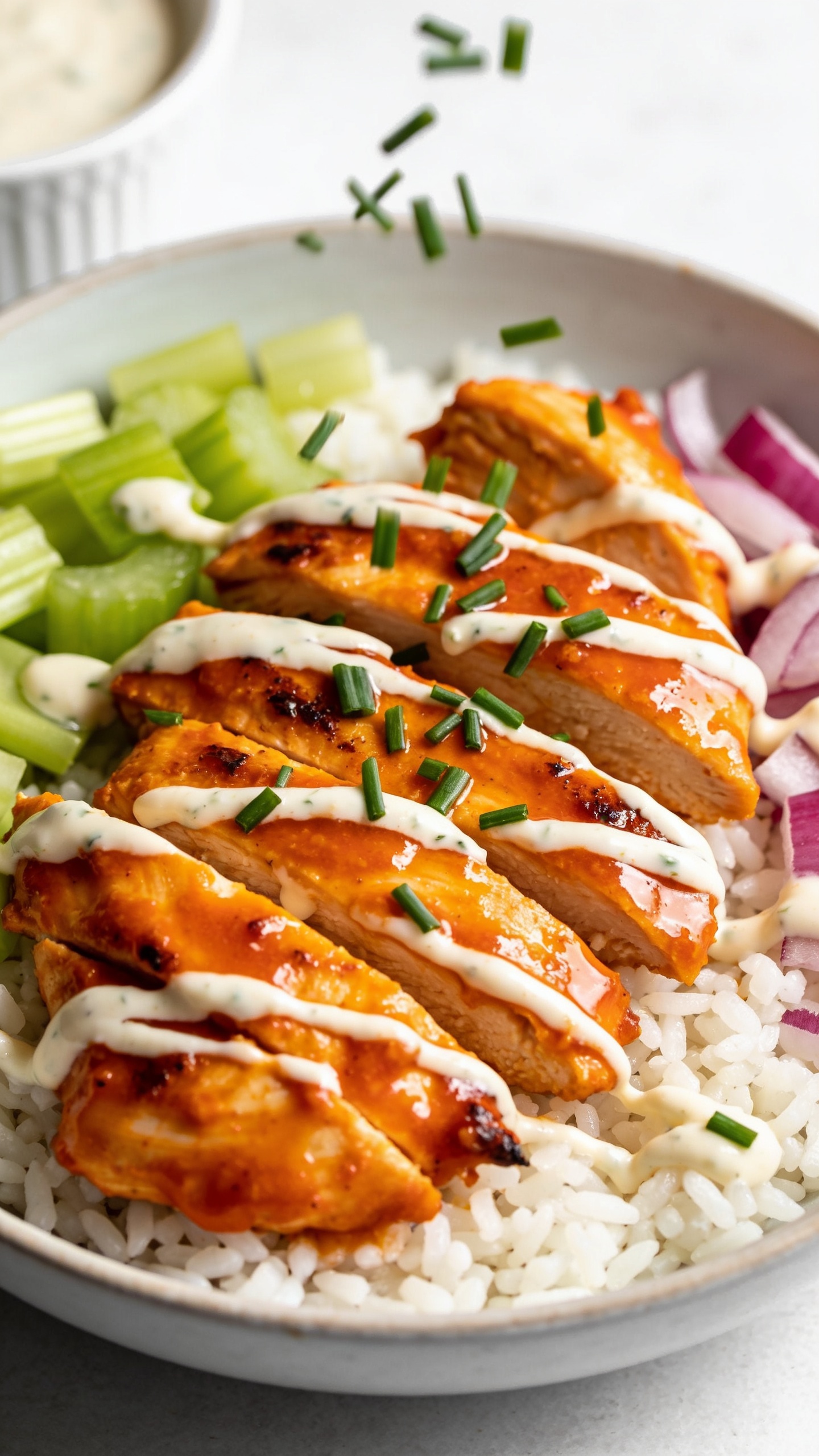 Tight macro angle from slightly overhead: consistent bowl of Buffalo ranch chicken over white rice with crisp celery and red onion, chicken slices fanned and lacquered in vibrant Buffalo sauce; delicate ranch drizzle added last, tiny droplets beading; fresh chives sprinkled mid-fall, some caught in motion above the chicken; edges of chicken glistening with a light char; grains of rice visible and fluffy; small ramekin of ranch barely cropped in the corner; bright natural light, shallow depth of field, rich textures and vibrant color contrast, food dominating 90â95% of frame.