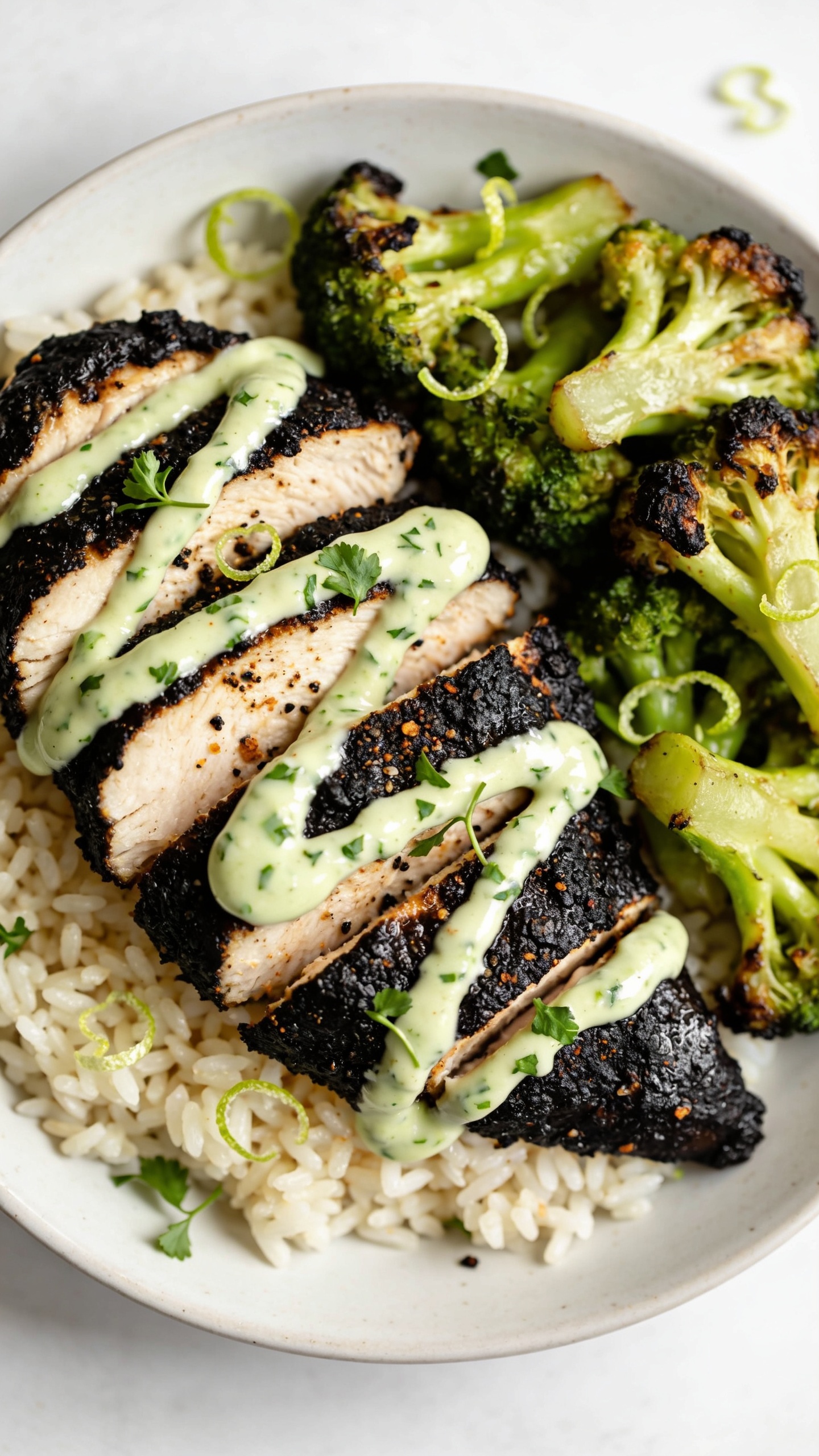 Ultra close-up angle from slightly overhead and to the side: consistent plated meal with sliced blackened chicken breast arranged over jasmine rice, roasted broccoli tucked alongside, and a glossy drizzle of cilantro-lime yogurt sauce zigzagging across the chicken. Emphasize crispy blackened edges, glistening surface, tender interior grain of the chicken, charred broccoli tips, flecks of coarse black pepper and smoked paprika visible on the crust. Finishing touches: micro-cilantro and tiny lime zest curls scattered. Bright natural light, shallow depth of field, 90â95% of frame filled with food, clean neutral backdrop, premium food-blog styling.