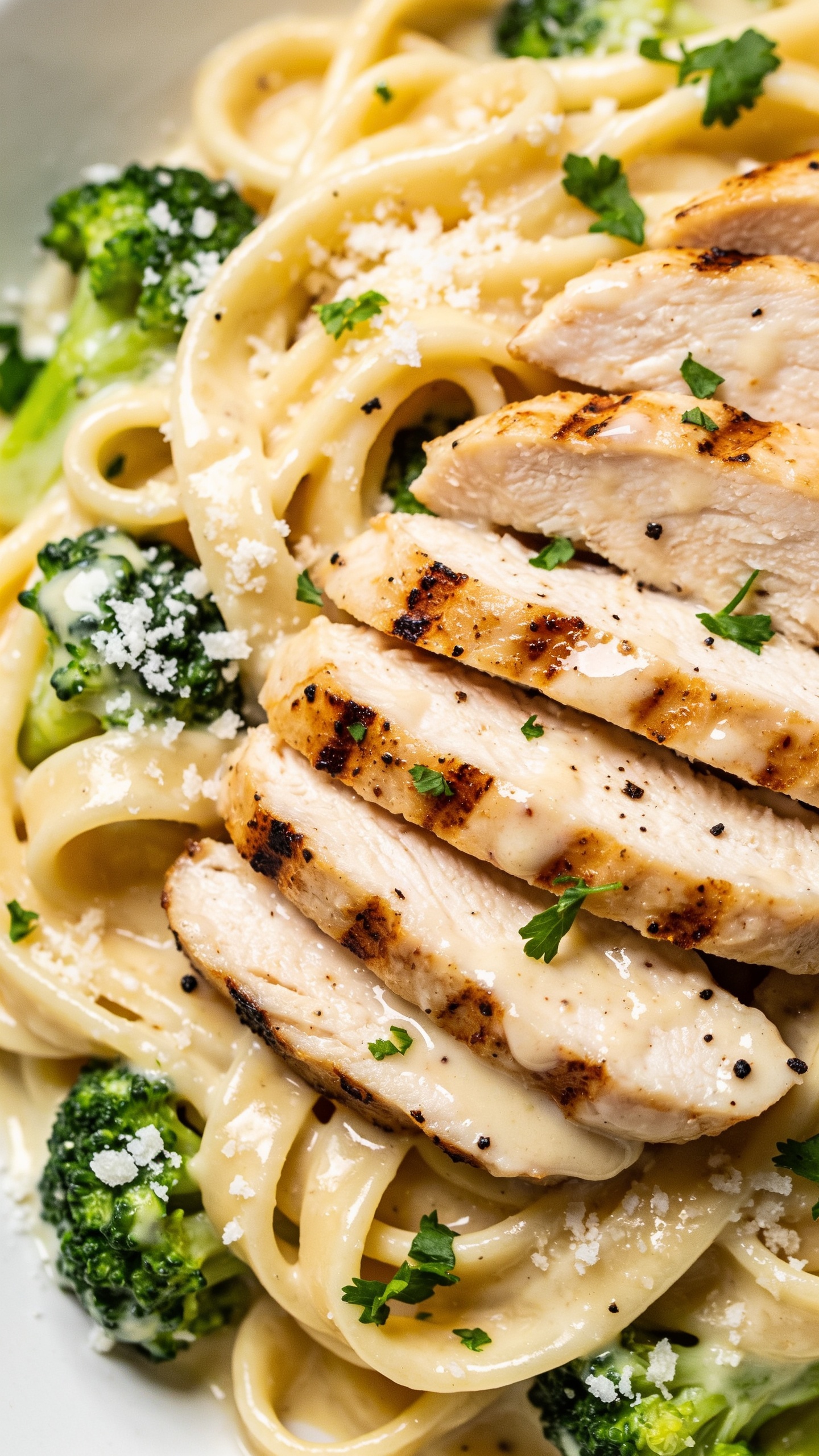 Extreme close-up angle on the plated dish from slightly overhead: dense nest of fettuccine lacquered in creamy light Alfredo with visible glossy sheen, several uniform slices of grilled chicken breast fanned over the pasta, edges lightly charred and juicy, broccoli florets nestled between noodles for vibrant green contrast. Freshly grated Parmesan scattered like snow, fresh parsley sprinkled, visible cracked black pepper. Bright natural lighting, shallow depth of field focusing on sauce texture and chicken juiciness, food occupying 90â95% of the frame, clean minimal background, no text.