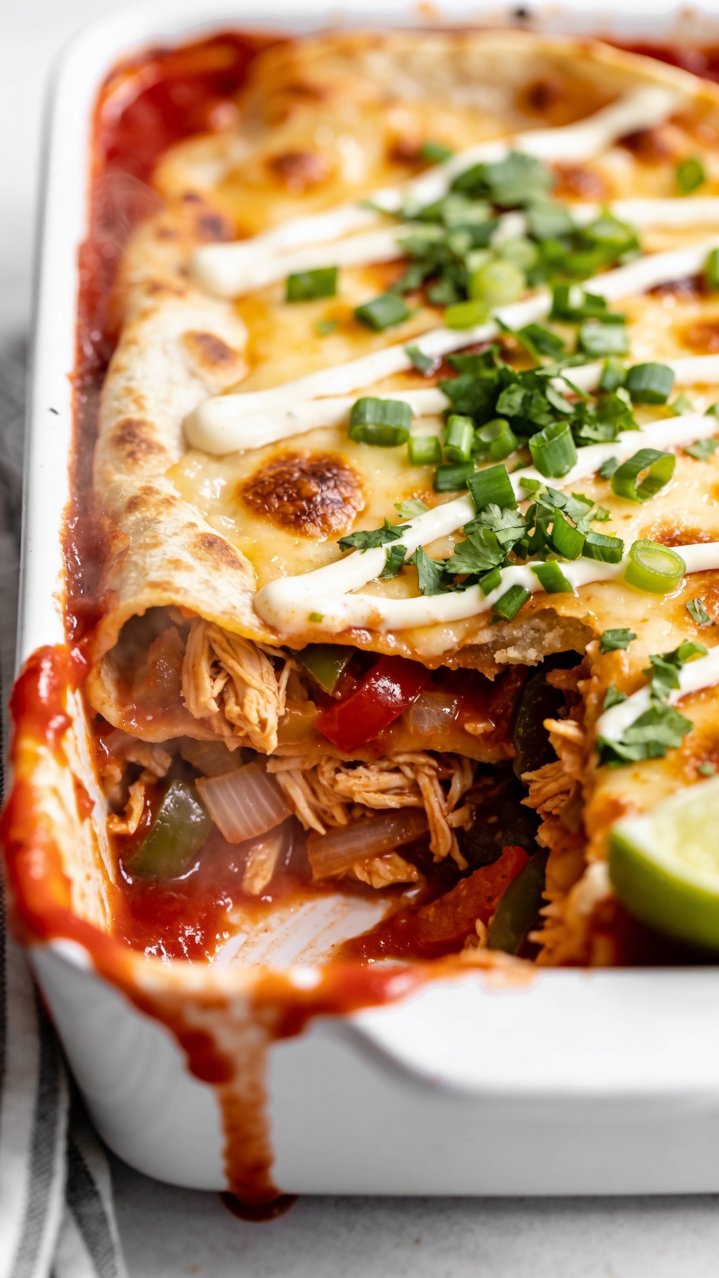 Macro close-up from a low 3/4 angle of the same finished chicken enchilada casserole in a small white meal-prep container, one corner scooped to reveal the interior layers. Focus on glossy red sauce pooling at the edge, crisp tortilla edges, juicy shredded chicken strands, sautĂŠed peppers and onions tucked between layers, and a molten cheese cap with browned blisters. Garnished with a tight sprinkle of chopped cilantro and green onion, a thin drizzle of crema zigzagging lightly, and a lime wedge peeking at the corner. Bright natural sidelight, shallow depth of field emphasizing the saucy cut edge, vibrant colors, steam faintly visible, food filling 90â95% of the frame, premium food-blog styling.