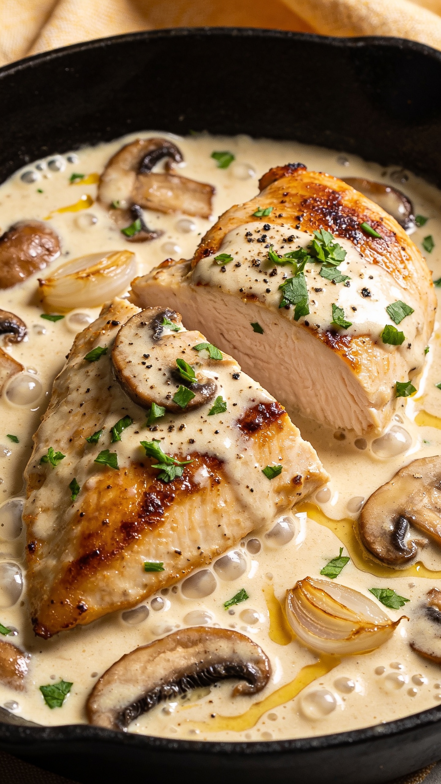 Extreme close-up angle from skillet edge: focus on one chicken breast partially sliced to reveal juicy interior, edges caramelized, blanketed in velvety cream sauce with browned mushroom slices and translucent sautéed garlic. A light scatter of fresh chopped parsley and a twist of black pepper on top, tiny bubbles and glossy ripples in the sauce, with golden fond streaks visible. Bright natural light, shallow depth of field, warm linen hint in background, 90–95% frame filled with the same finished dish, cohesive premium styling.
