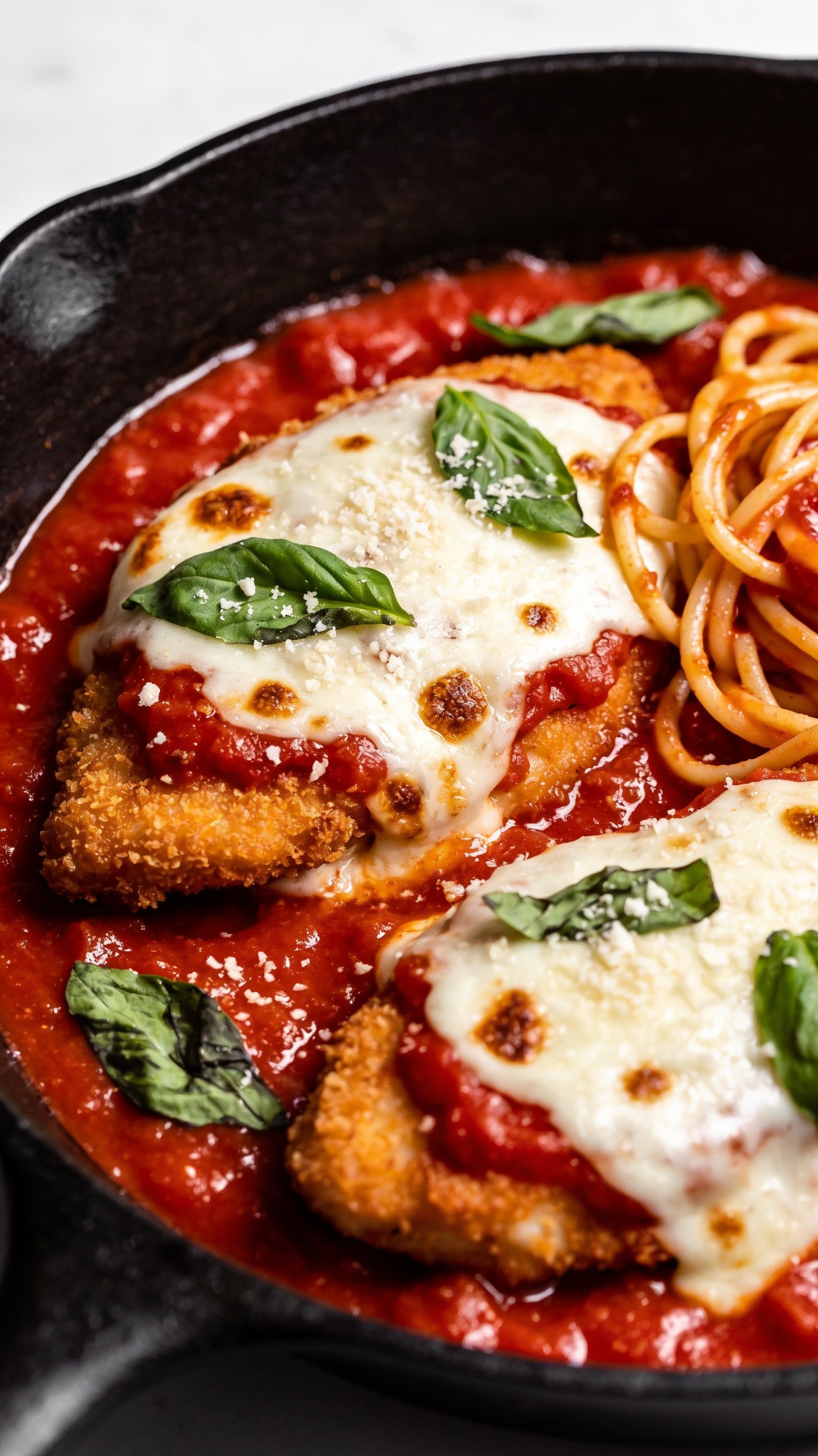 Ultra close-up hero shot of One Pan Chicken Parmesan in a black cast-iron skillet: golden, crispy breaded chicken cutlets nestled in glossy, chunky marinara, crowned with melted mozzarella and a light sprinkle of freshly grated parmesan and torn basil leaves. Edges of cheese lightly blistered with browned spots, cutlets partially submerged so the crust remains visibly crisp above the sauce. A few spaghetti strands twirled at the side of the skillet, lightly coated in marinara for context. Bright natural side lighting, shallow depth of field, food filling 90â95% of frame, vibrant reds, creamy whites, golden crust, and fresh green basil, premium food-blog styling.