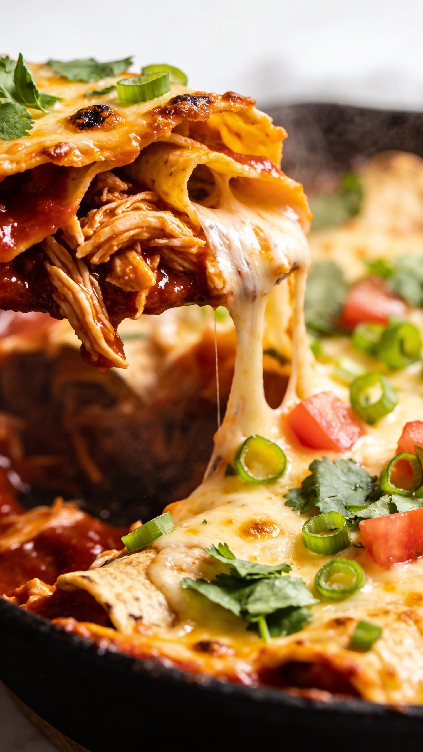 Macro close-up of a scooped section in the skillet revealing the inside layers of the same chicken enchilada bake. The foreground showcases juicy shreds of chicken saturated in deep red enchilada sauce, ribbons of toasted corn tortillas, and glossy, stringy melted cheese pooling and stretching into the cavity. Caramelized cheese crust along the rim, tiny charred cheese blisters, and saucy sheen throughout. Fresh cilantro leaves and green onion rounds sprinkled over the top, a few tomato dice nestled into the cheese. Bright natural top-left lighting, shallow depth of field, steam gently visible, rich textures and saturated color, food filling the frame for a premium food-blog look.