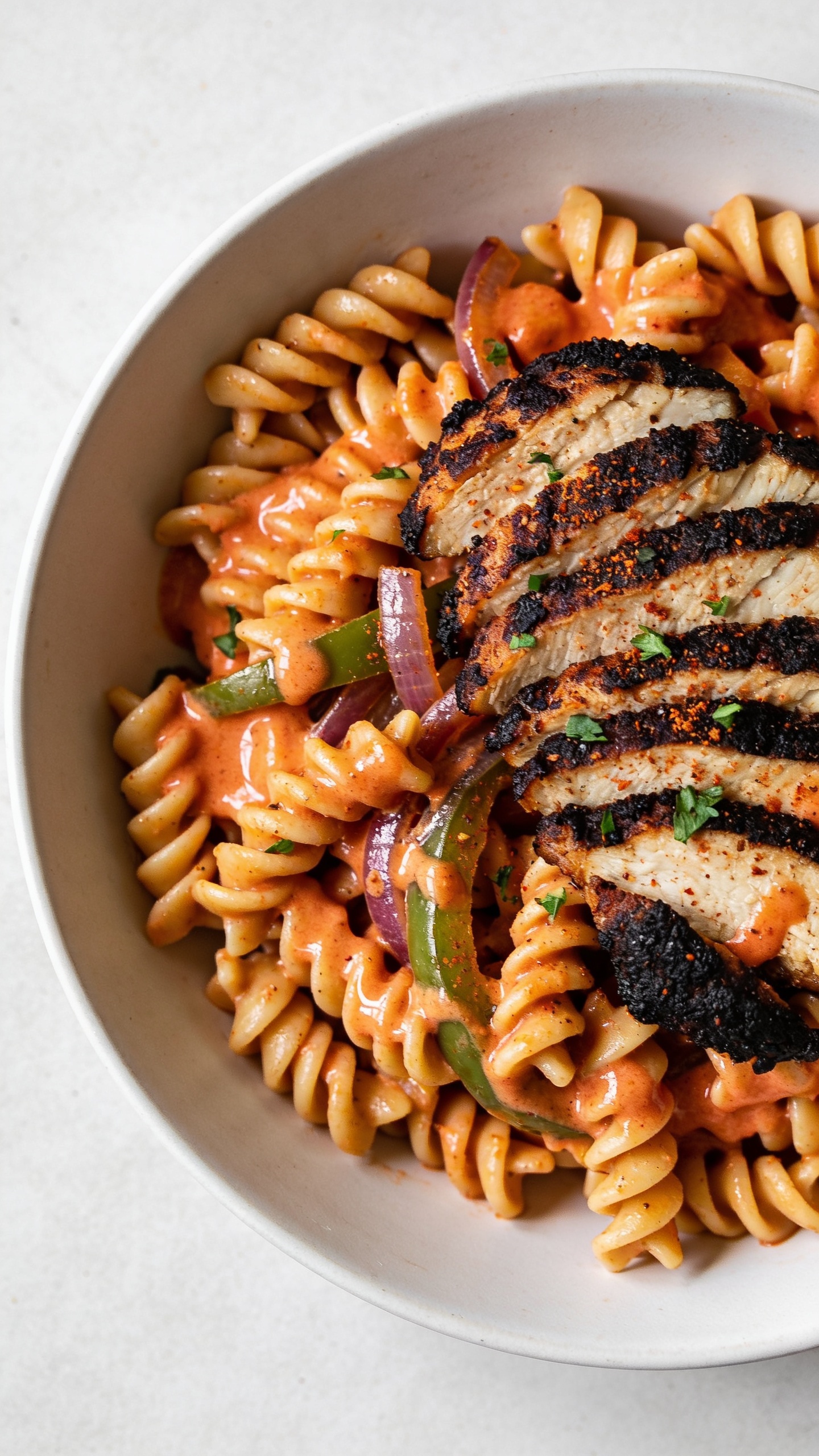 Macro angle from slightly overhead: the same Healthy Cajun Chicken Pasta in a matte white shallow bowl, dense composition with pasta spiraled toward the center; creamy orange-red Cajun yogurt sauce glistening, collecting in glossy pools between penne; blackened chicken slices fanned on top showing juicy interior and charred spice crust; sautĂŠed bell pepper and onion strips tucked in; a delicate final dusting of Cajun seasoning and finely chopped parsley visible on the surface; bright natural side light, shallow depth of field emphasizing saucy ridges and textures; neutral, uncluttered background; food fills 90â95% of frame, consistent with the hero styling.