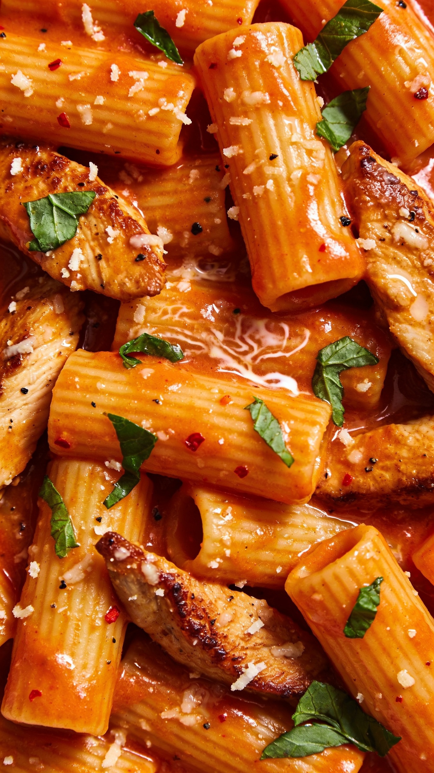 Macro-level overhead-angled close-up of the same finished dish: rigatoni densely coated in a rich, orange-red creamy tomato sauce with silky shine, browned chicken slices peeking through, finely grated parmesan melting into tiny glossy pools, fresh basil torn into small pieces scattered across. Flecks of black pepper and red pepper flakes visible, slight swirl pattern in the sauce showing cream emulsification. Bright natural lighting, shallow depth of field emphasizing saucy ridges and crispy chicken edges, food filling 90â95% of frame, cohesive with the other images.