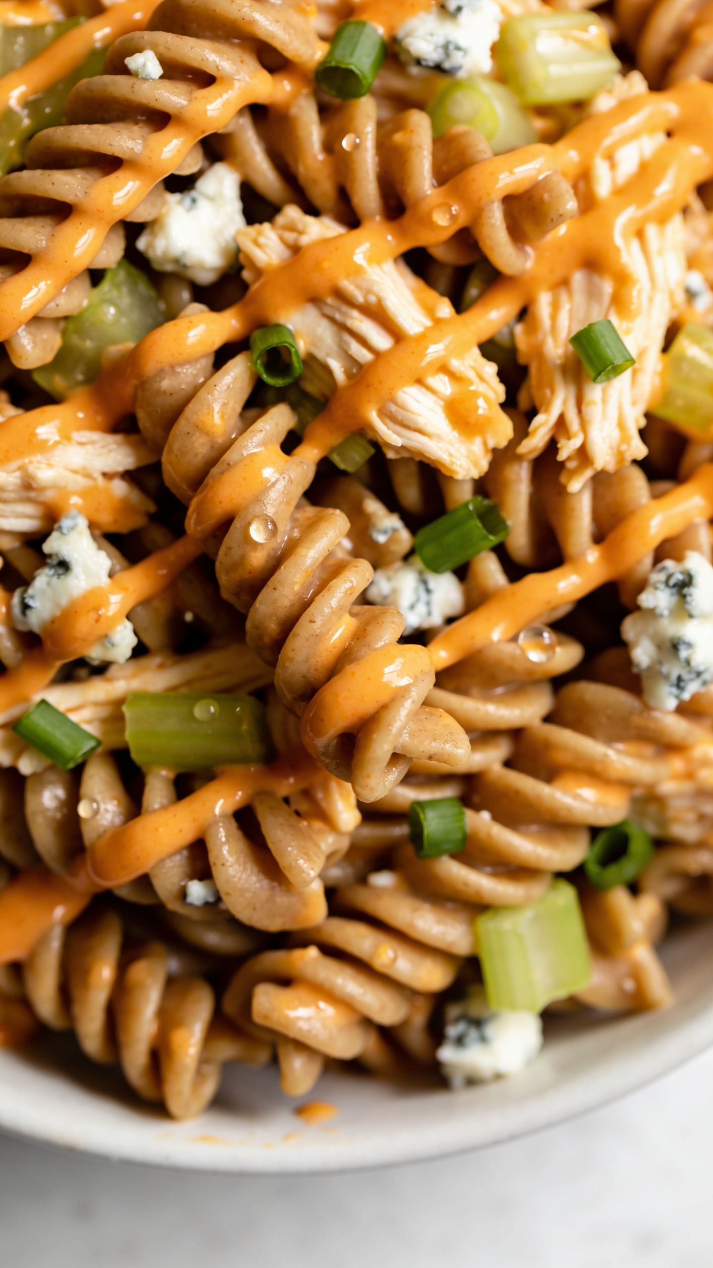 Ultra close-up overhead-angled detail of the same finished Healthy Buffalo Chicken Pasta, emphasizing textures: curls of whole-grain rotini coated in creamy orange sauce pooling lightly in the grooves, visible moist shreds of chicken, tiny blue cheese crumbles just beginning to soften, crisp celery bits and vibrant green onions scattered. A glossy drizzle of buffalo sauce zigzagged very lightly for sheen, micro droplets catching the light. Bright natural lighting, shallow depth of field isolating a small cluster of pasta in razor focus, minimal neutral background, food dominating 90â95% of the frame, clean modern styling.