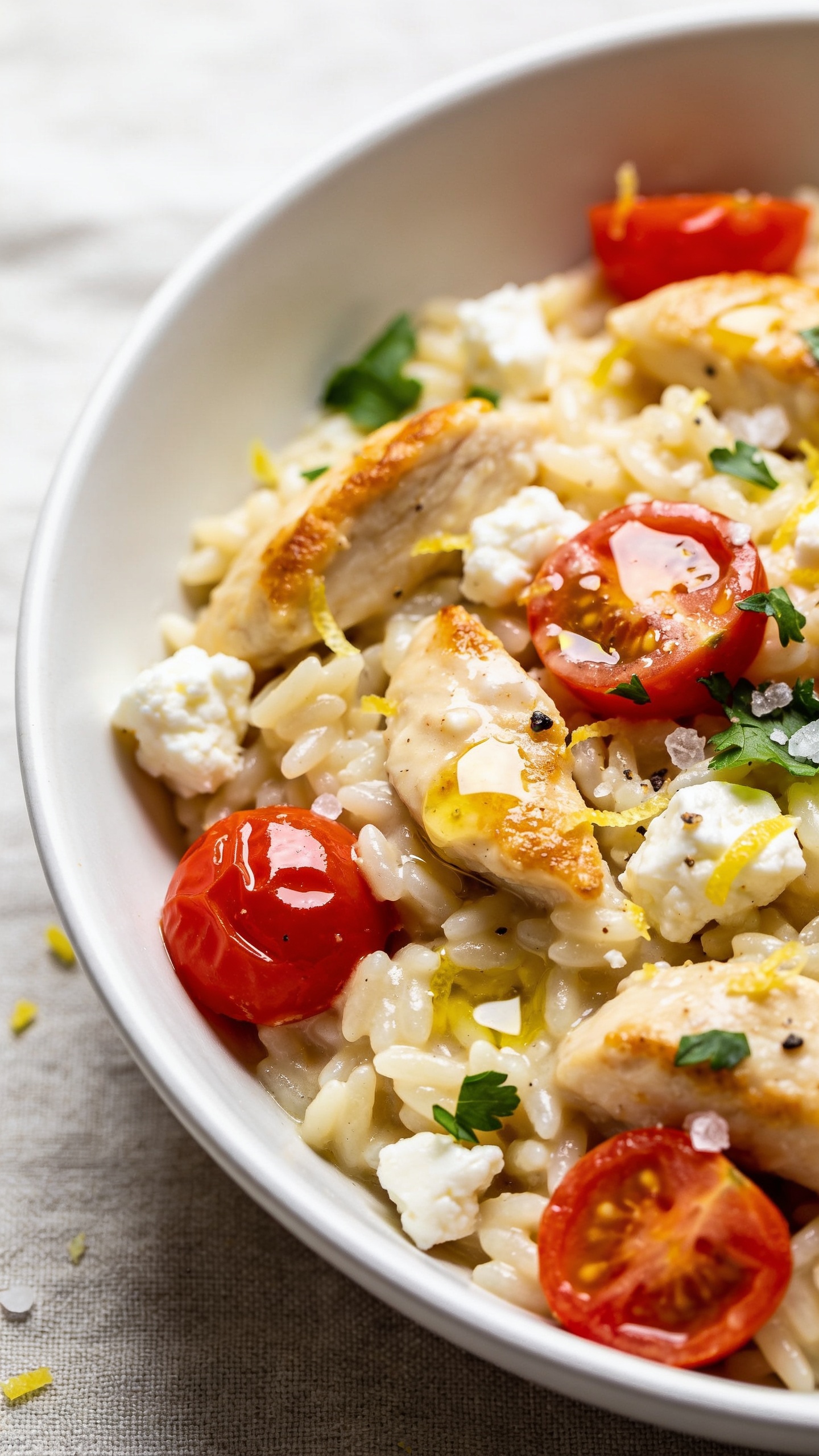 Ultra close-up angled top-down detail of the same finished recipe plated in a shallow white bowl for variety but identical styling: orzo creamy and silky, chicken pieces golden at the edges, feta crumbles gently softened and marbled through, cherry tomatoes glossy and just-split, micro-puddles of lemony olive oil, fine lemon zest scattered, and fresh parsley leaves. A few coarse salt flakes and cracked pepper visible. Vibrant colors, shallow depth of field emphasizing juicy textures and glossy sauce, bright natural light from the left, neutral linen surface, premium food-blog look. No text, no hands.