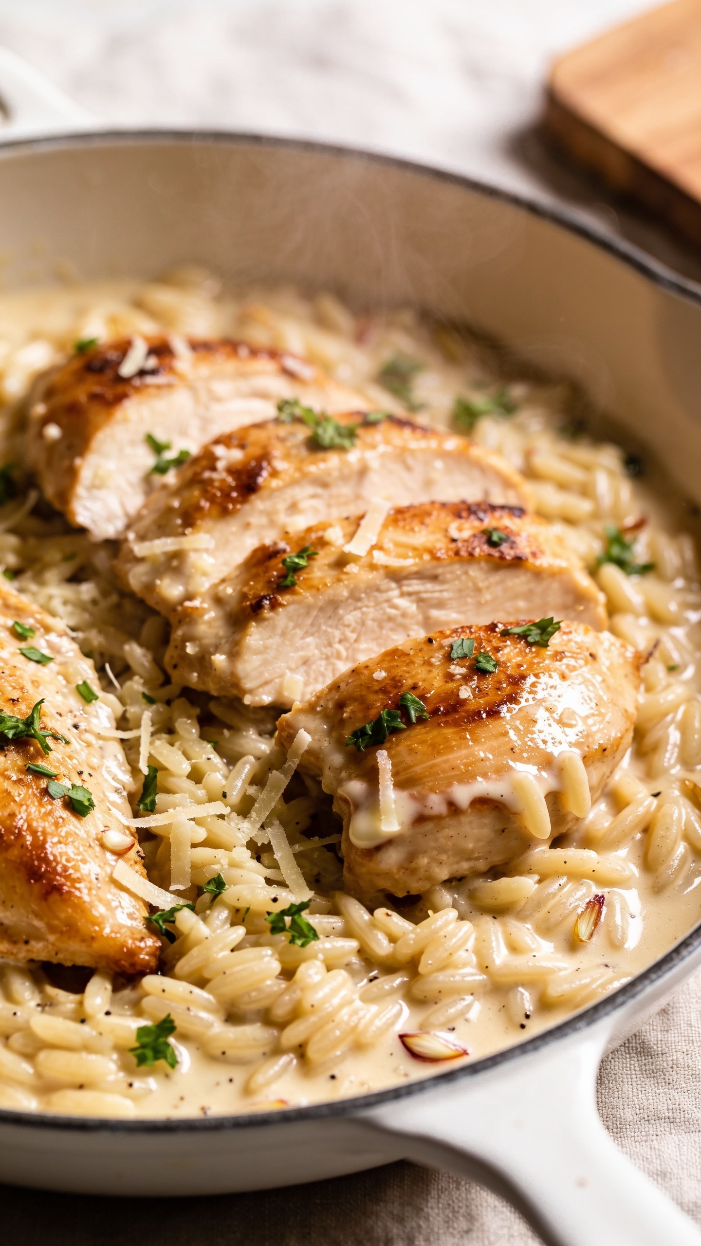 Ultra-close hero shot of Creamy Garlic Chicken Orzo in a shallow white skillet, food filling 90â95% of frame. Golden seared chicken thigh slices nestled into glossy, creamy garlic-Parmesan sauce with tender orzo, tiny flecks of sautĂŠed garlic visible, micro-shavings of Parmesan melting at the edges, and a sprinkle of freshly chopped parsley. Orzo grains plump and coated, sauce silky with gentle sheen, light pepper specks. Bright natural side lighting, shallow depth of field, softly blurred neutral linen and a hint of wooden board in the background. Steam subtly rising, textures crisp and creamy, premium food-blog styling, consistent color palette of warm golds, ivory sauce, and fresh green herbs.