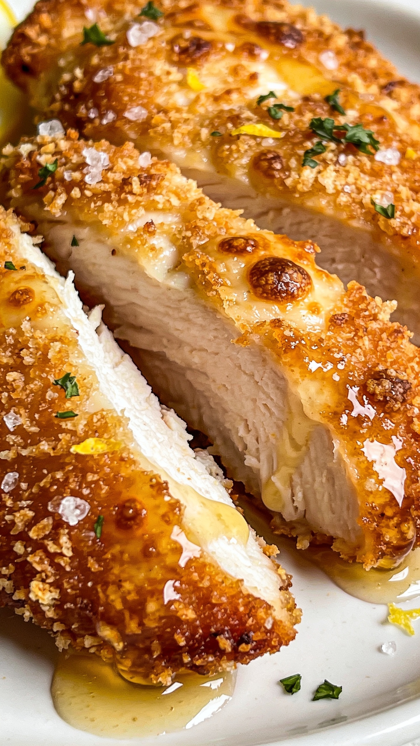 Macro close-up from a different angle focusing on texture: a sliced cutlet showing the juicy white chicken inside contrasted with the deeply golden, crackly Parmesan-panko crust; tiny browned cheese bubbles, crisp edges, and coarse sea salt crystals visible; glossy drizzle trails of garlic-butter pooling lightly, with micro parsley scattered; a faint zest dusting from lemon visible on the crust; same plate and setting for continuity; bright natural light, shallow depth of field, 90â95% frame filled with the chicken, rich, crisp, irresistible styling.