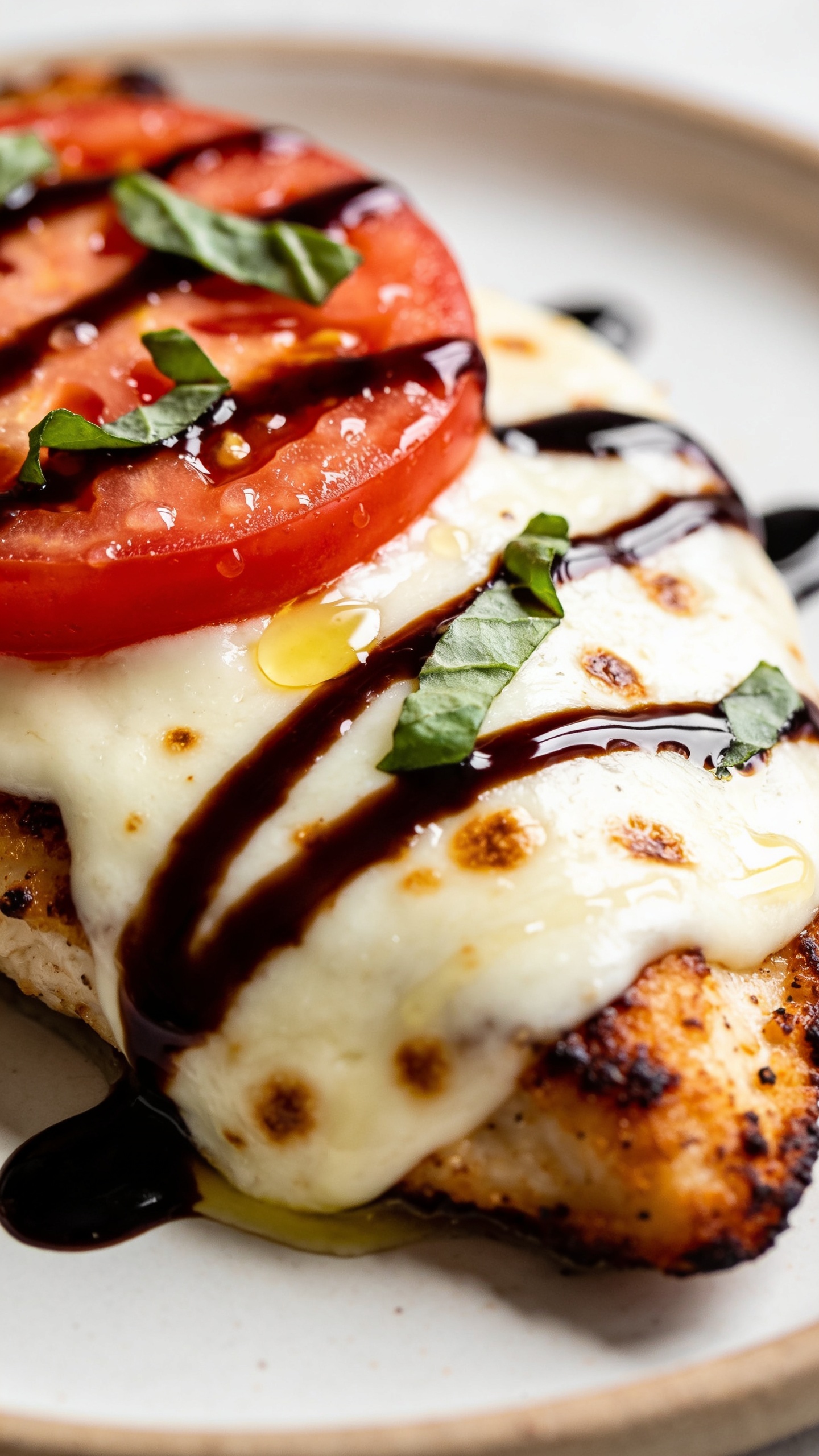 Macro angle from the side focusing on textures: the same finished Air Fryer Chicken Caprese on the plate, showing crispy seared edges of the chicken, mozzarella melted to a glossy blanket with browned speckles, juicy tomato slices beading with moisture, torn basil leaves scattered on top; a final slow-motion look of thick balsamic glaze already settled in shiny swirls across the cheese, tiny olive oil sheen catching the light; bright natural lighting, shallow depth of field emphasizing the caramelized rim and melty cheese, 90–95% food in frame, clean modern styling with minimal neutral background blur.
