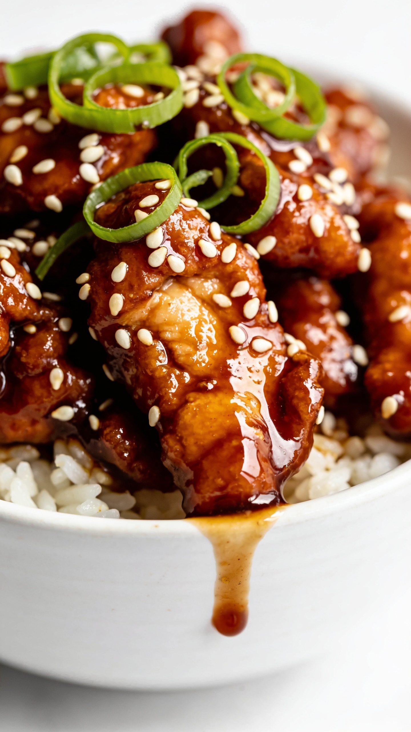 Macro angle from slightly overhead and to the side: Air Fryer Sesame Chicken tossed in a thick, shiny sesame-ginger glaze, tightly framed to emphasize caramelized, craggy edges and juicy interiors, with sesame seeds clinging to the sauce and fresh green onion curls across the top. A small glossy puddle of sauce glints at the rim of the white bowl, with fluffy rice peeking from underneath. Bright natural light, shallow depth of field, 90â95% food in frame, cohesive with the hero image, clean minimal background, premium, craveable texture focus.