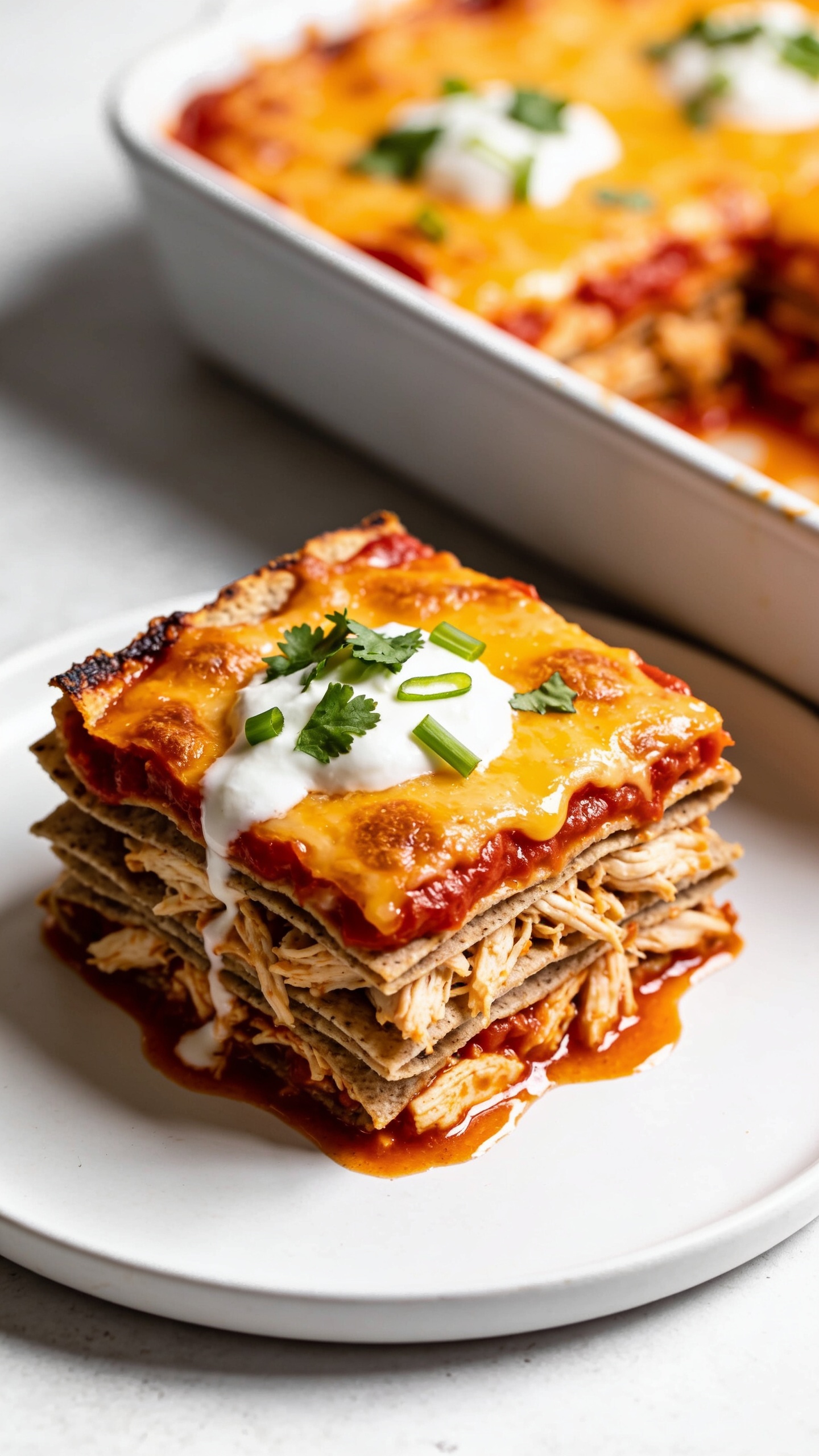 Tight angled close-up slice view plated on a simple white plate beside the baking dish: a neatly cut square of Healthy Chicken Enchilada Casserole showing clean, stacked layers of whole-grain tortillas, tender shredded chicken, and vibrant red enchilada sauce, crowned with a molten, lightly browned cheese cap; small dollops of Greek yogurt softly melting into the top, sprinkled with chopped cilantro and a few thin green onion slices; glossy sauce pooling slightly at the base, crispy edges visible; bright natural light, shallow depth of field, minimal props, vibrant yet clean premium food-blog styling.