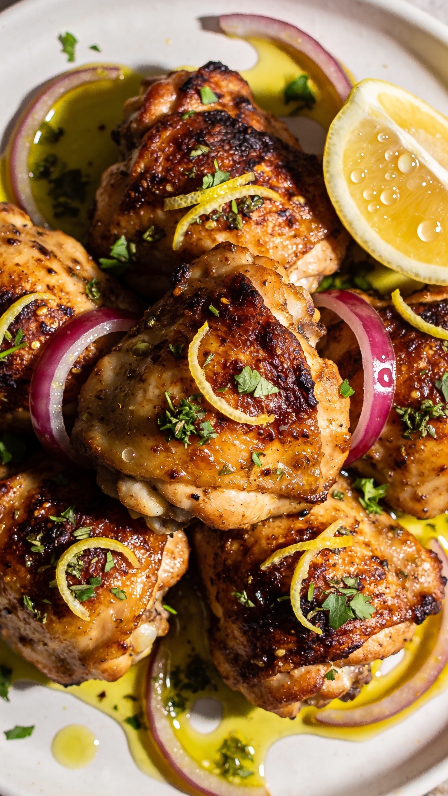 Macro angle from slightly overhead: tightly packed cluster of the same Greek chicken thighs with intensely crisp, browned skin, tiny blistered bubbles, and charred edges; zesty detail of fresh lemon zest sprinkled over, plus chopped oregano and parsley; glossy olive oilâlemon-garlic sauce pooled in spoonable rivulets around the thighs; a couple of thinly sliced red onions softened and glistening; a wedge of lemon squeezed, with visible juice droplets beading on the skin; bright natural light raking across to highlight crunch and juiciness; shallow depth of field; 90â95% of the frame filled with the finished dish on a simple white plate.