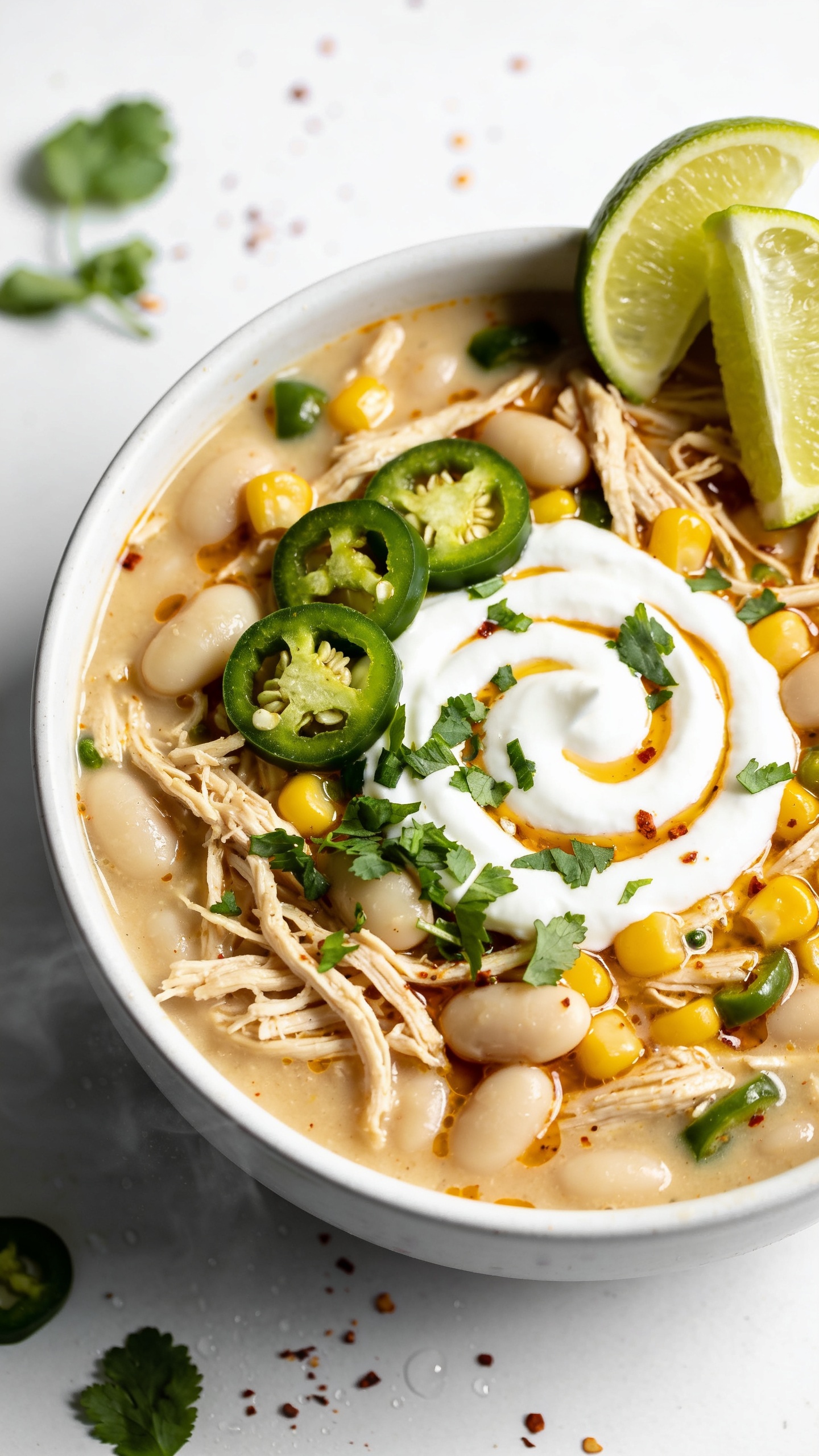 Macro overhead three-quarter angle of the finished chili in the same matte white bowl, emphasizing texture: creamy broth pooling around shredded chicken fibers, white beans, and corn, with tiny green chile flecks and a light chili oil sheen. Identical garnishes for continuityâGreek yogurt swirl centered, chopped cilantro scattered, precise jalapeño rounds, lime wedge on rim. A few extra cilantro leaves and a pinch of chili flakes artfully on the surface. Bright natural daylight, shallow depth of field, subtle condensation and soft steam visible, food occupying 90â95% of the frame, clean minimal backdrop for a high-end food-blog look.