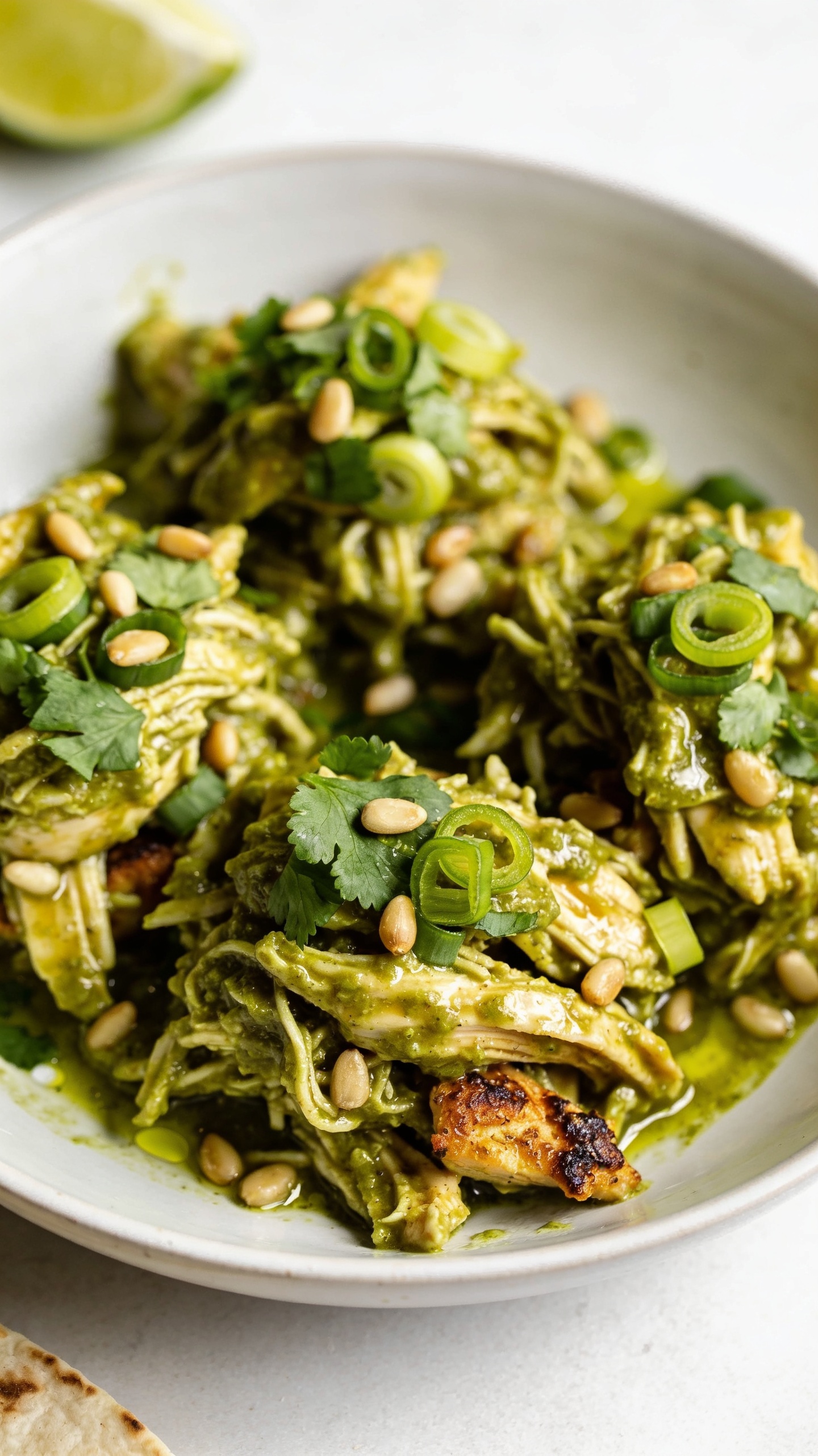 Tight overhead three-quarter close-up of the same finished salsa verde chicken in a shallow white bowl: richly sauced shreds arranged in natural mounds, glistening with a light lime sheen; garnished identically with chopped cilantro, green onion curls, and a few toasted pepitas; tiny pools of salsa verde collecting at the edges, crispy caramelized bits of chicken peeking through; a lime wedge and a corner of a tortilla slightly out of focus at the edge; bright natural light, shallow depth of field, rich textures and vibrant green-gold palette, food filling 90â95% of the frame.