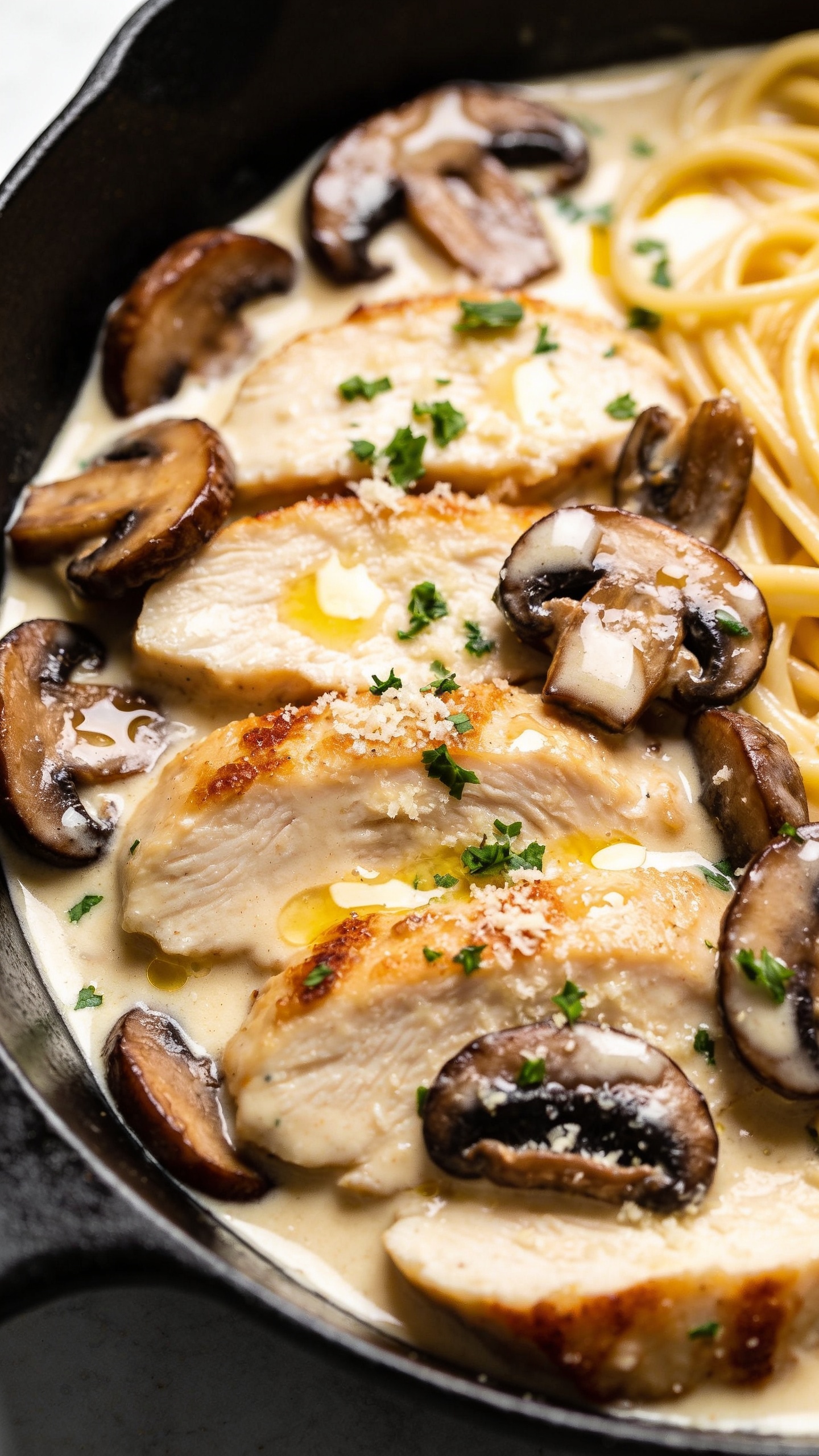 Tight overhead three-quarter close-up of the same finished dish in the black skillet: chicken slices nestled among deeply browned mushrooms, all lacquered with a luscious ivory-gold cream sauce; tiny pools of sauce glinting with butter, edges of chicken slightly crisp, mushrooms glossy; a light sprinkle of chopped parsley and a whisper of grated parmesan melting into the cream; a few egg noodles swirling under the sauce for contrast; bright natural lighting, shallow depth of field emphasizing juicy textures and creamy sheen, food filling 90–95% of the frame with cohesive premium food-blog styling.
