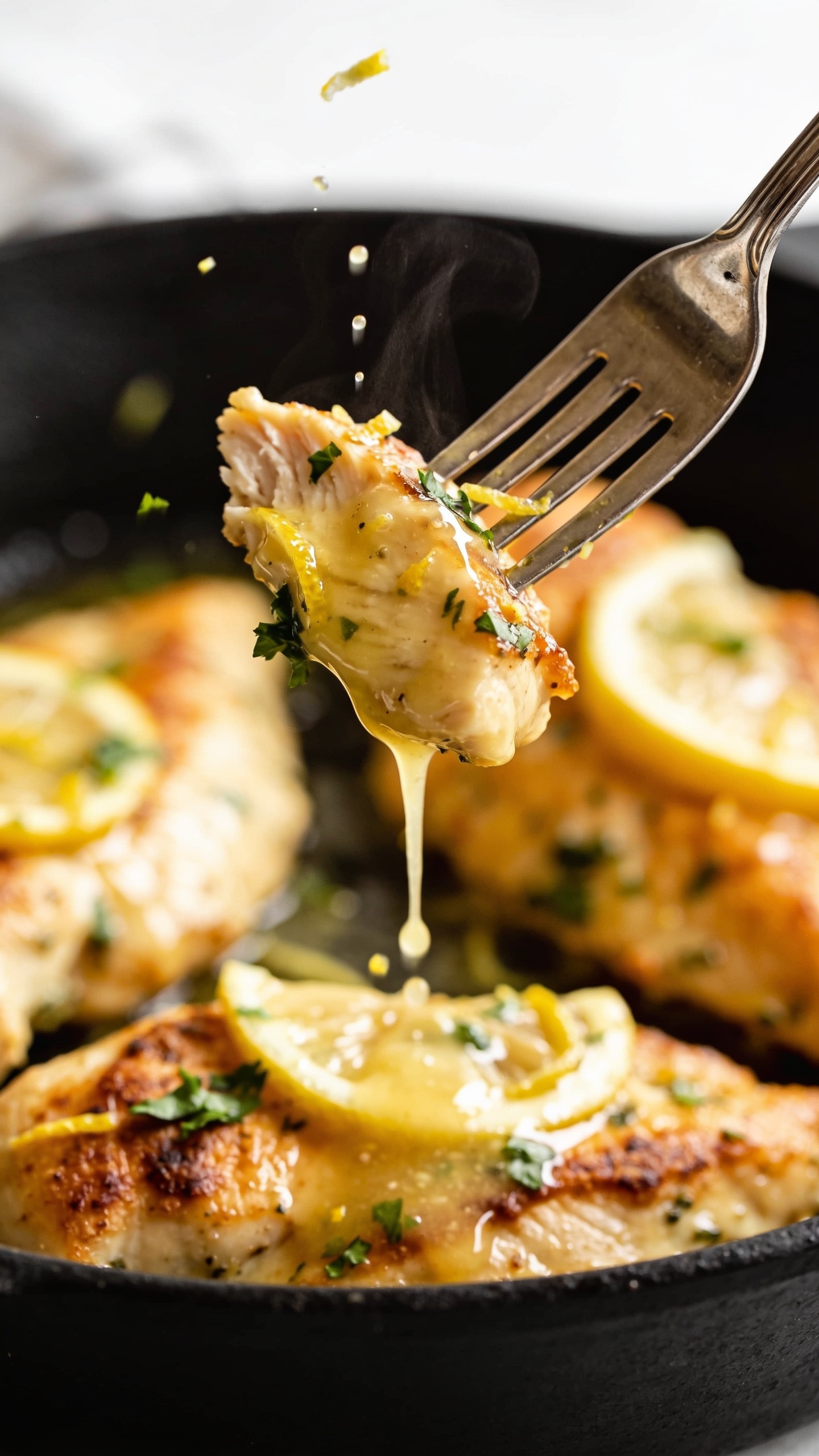 Dynamic action close-up: a polished dinner fork lifting the perfect bite-sized piece of the same lemon-herb chicken from the skillet, the bite glistening with glossy lemon-butter sauce; a silky ribbon of sauce trailing back to the skillet, tiny droplets suspended midair; flecks of lemon zest and fresh parsley clinging to the bite, steam subtly rising; the remaining chicken in the skillet blurred but clearly golden with crispy edges and thin lemon slices peeking through; bright natural light, shallow depth of field, intense texture and moisture, no hands visible, food fills 90â95% of frame.