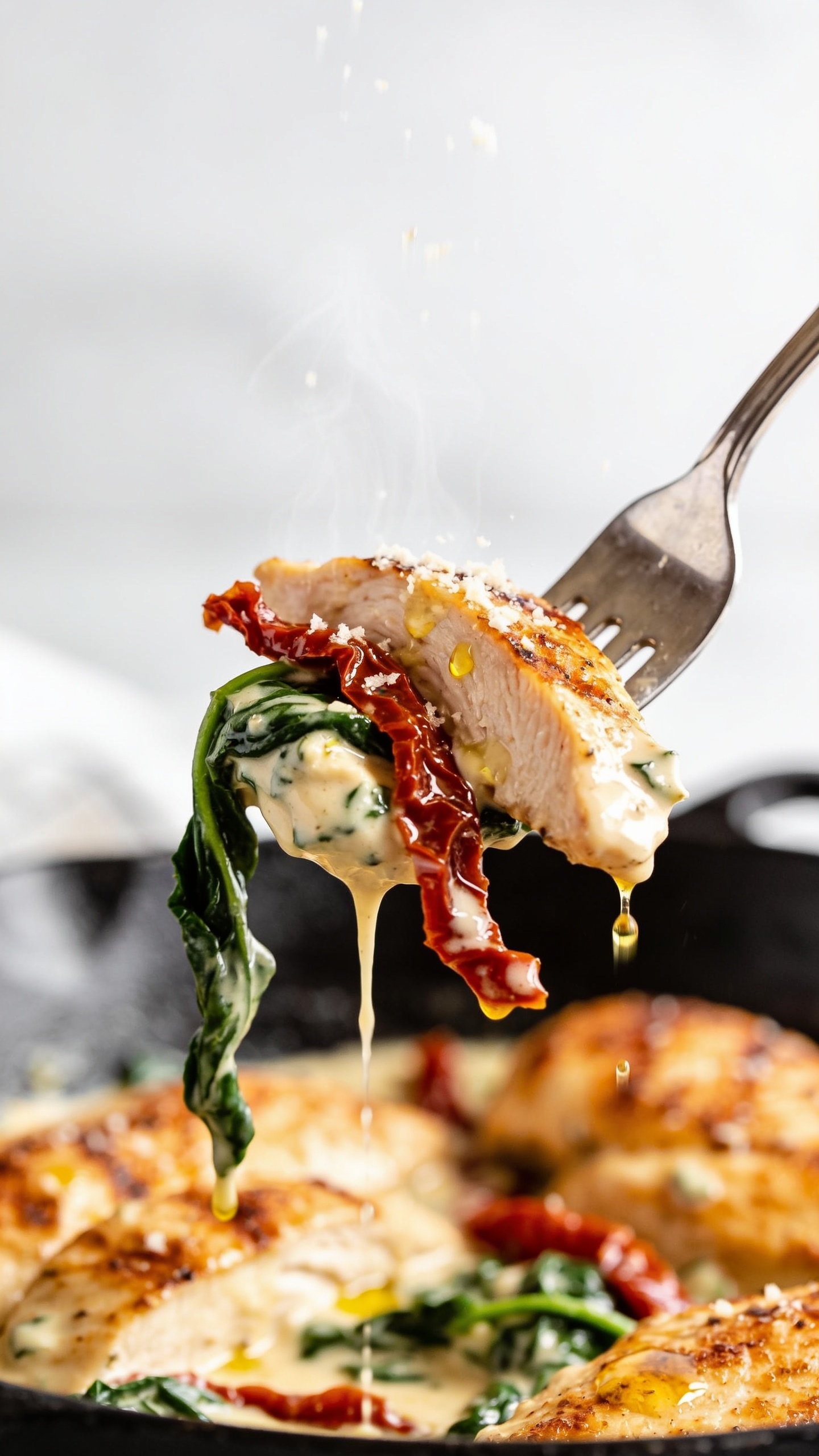 Dynamic bite moment: a stainless fork lifting the perfect bite from the skilletâone juicy slice of golden-seared chicken coated in creamy garlic sauce, a ribbon of sun-dried tomato draped over it, a strand of wilted spinach clinging, and a glossy drip of sauce slowly falling back into the skillet below. Only the fork visible, no hands. Steam subtly rising, bright natural light, shallow depth of field with the lifted bite in sharp focus and the same skillet of Sun-Dried Tomato Chicken Skillet blurred beneath. Parmesan flecks melting on the bite, micro droplets of olive oil shimmering. Food fills 90â95% of the frame, capturing movement and anticipation one second before the bite.