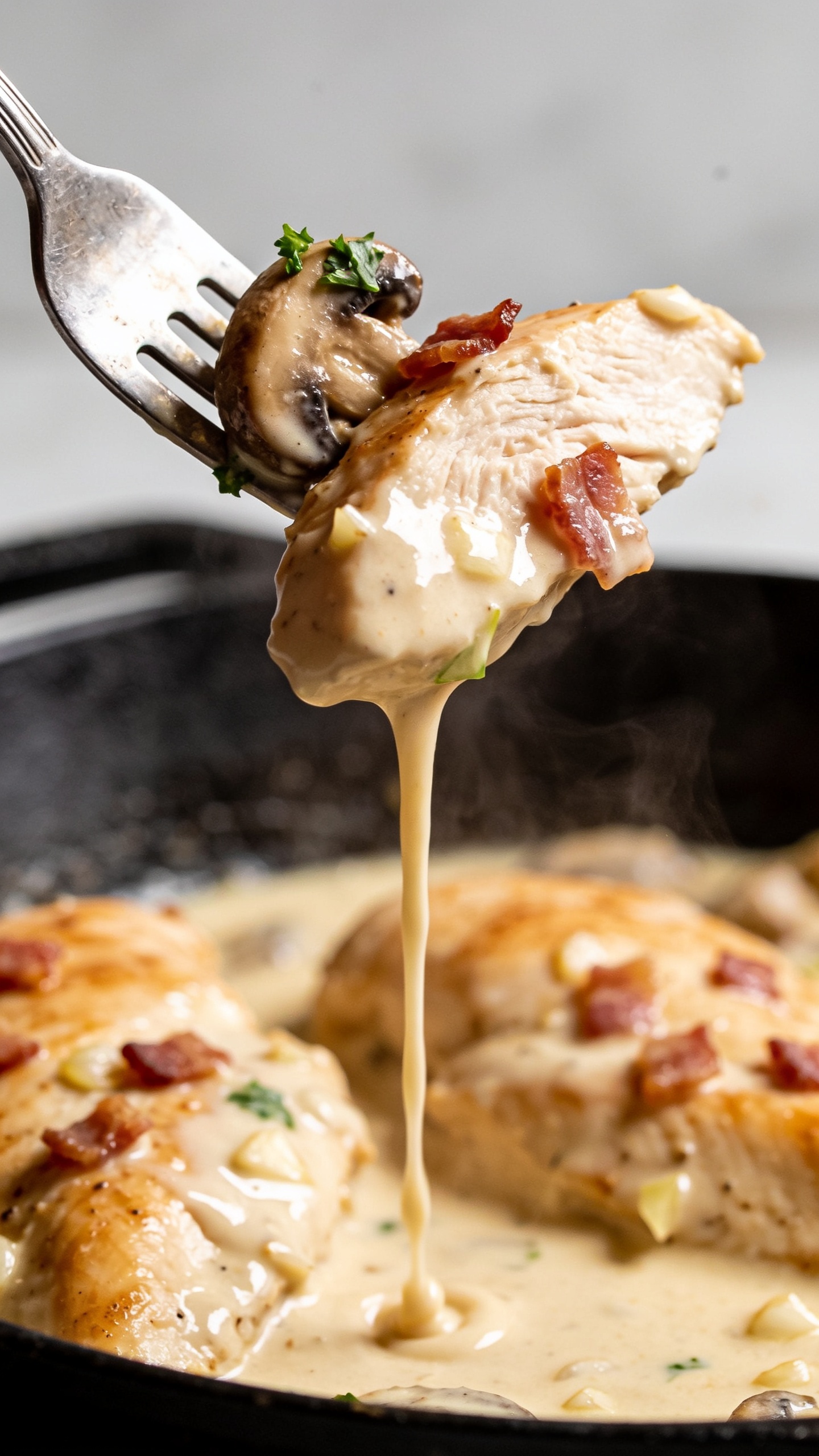 Dynamic action close-up capturing the irresistible bite: a fork lifting a perfect piece of the juicy chicken breast, coated in the glossy garlic-cream sauce, a ribbon of sauce slowly dripping back into the skillet below. Tiny bacon shards and a fleck of parsley cling to the bite, with a tender piece of sautĂŠed mushroom caught on the tine. The skillet with remaining chicken and creamy sauce is blurred in the background; bright natural lighting, shallow depth of field, visible steam, and rich highlights on the silky sauce. No hands or people, food dominates the frame.