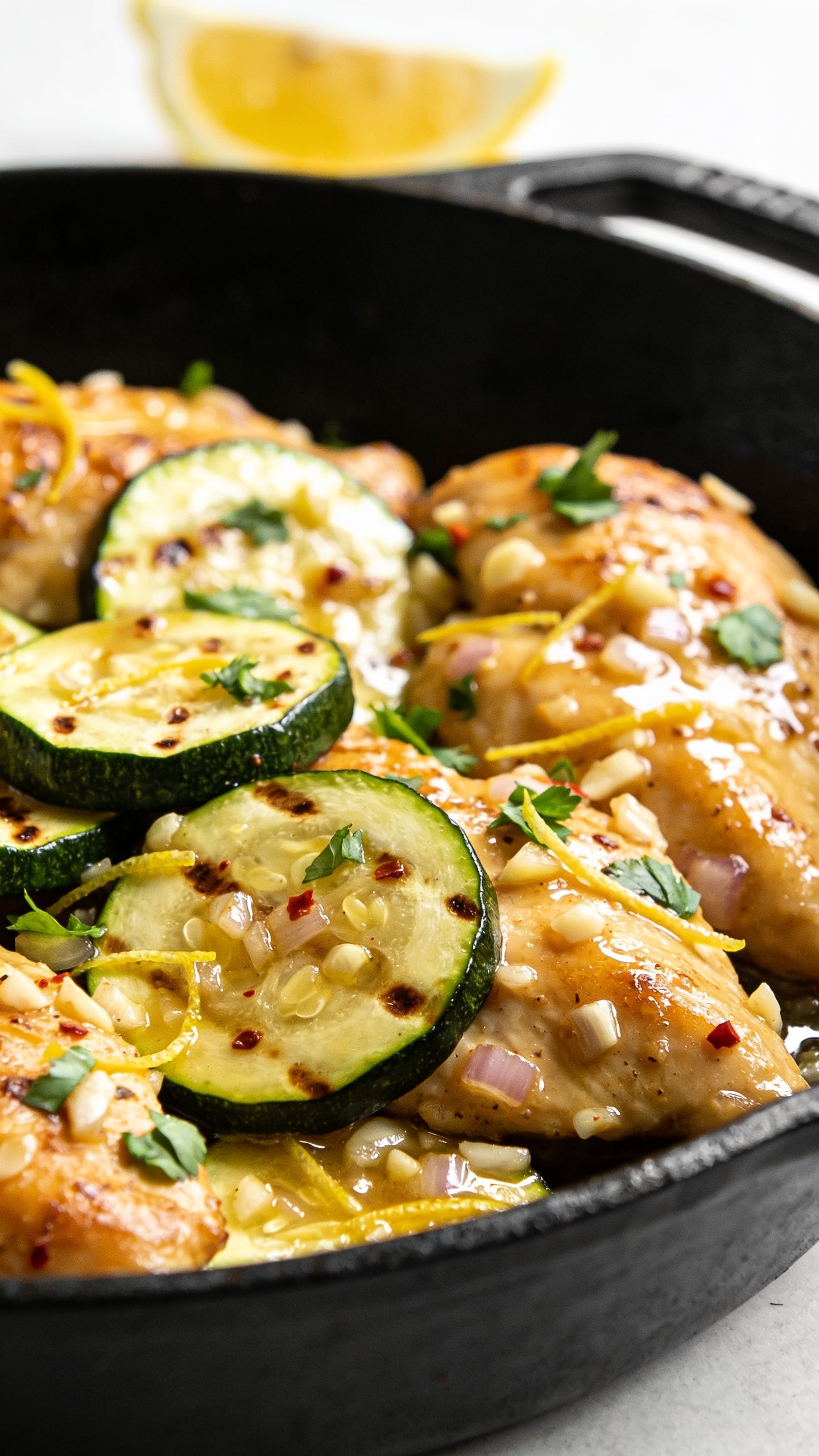Extreme close-up angle from skillet edge: focus on overlapping slices of tender-crisp zucchini with browned spots, nestled against succulent, golden-seared chicken pieces, all coated in a shiny garlic-lemon pan sauce with visible minced garlic and shallot bits; scattered fresh parsley, a whisper of red pepper flakes, and ultra-fine lemon zest strands. A thin wedge of lemon slightly out of focus in the background. Bright natural lighting, shallow depth of field, rich textures and vibrant colors, food filling 90–95% of the frame, same cast-iron skillet and styling as the hero for consistent look.