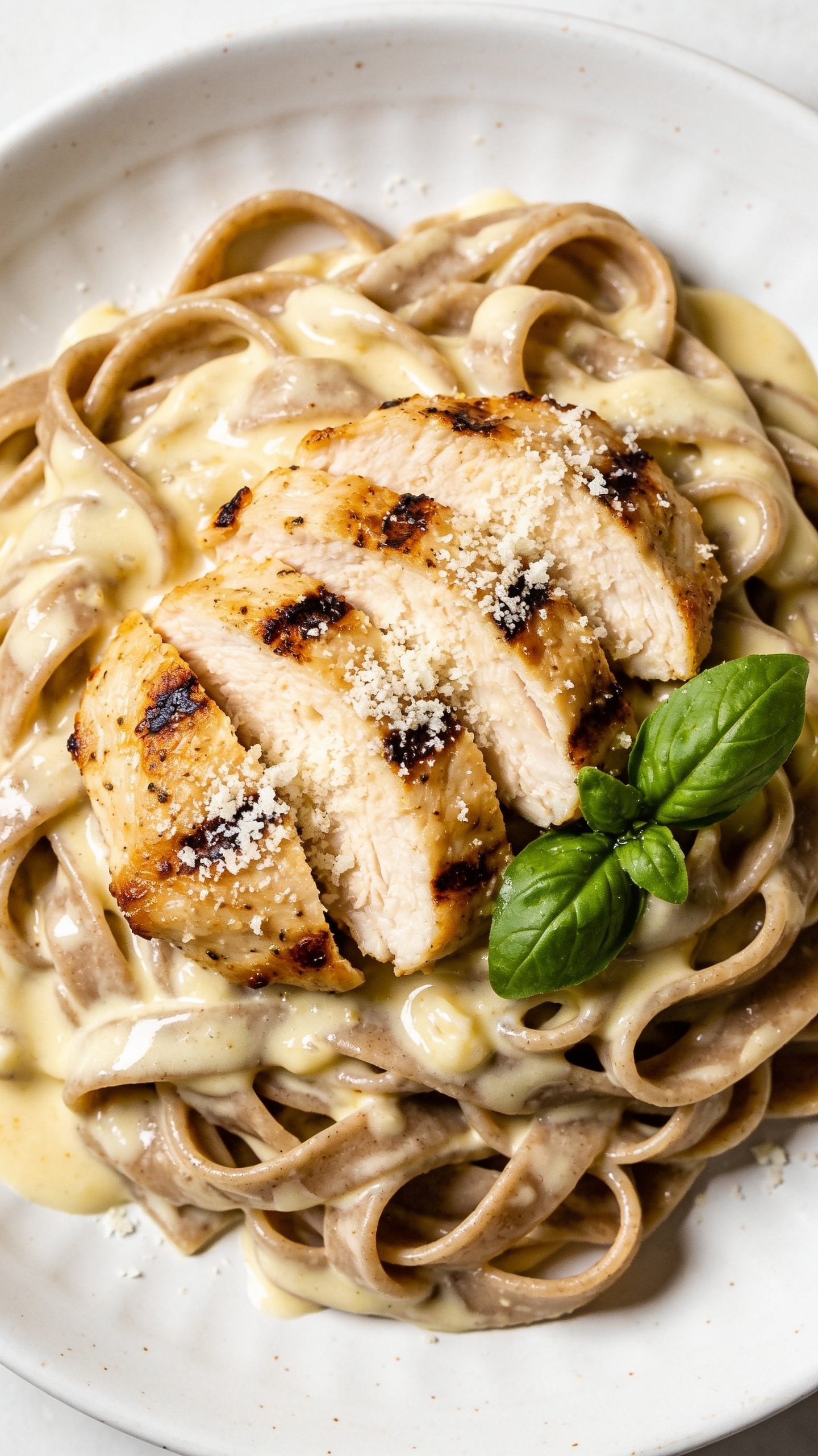 A stunning hero shot of the finished High Protein Grilled Chicken Alfredo Pasta plated on a white ceramic plate, glistening creamy Alfredo sauce coating perfectly cooked whole-wheat fettuccine, tender grilled chicken slices arranged atop, a light dusting of grated Parmesan, and a sprig of fresh basil; bright natural lighting, shallow depth of field, rich textures, no people, 90–95% frame filled with the dish.
